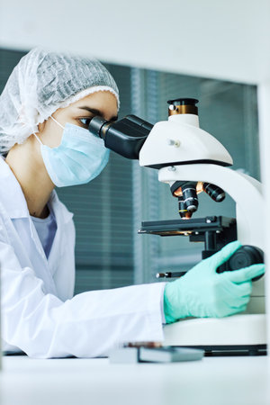 Vertical side view portrait of female scientist looking in microscope while working in medical laboratory, blue tonesの写真素材