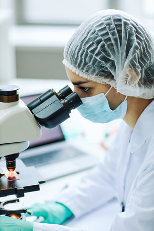 Vertical side view portrait of young female technician looking in microscope while doing research
