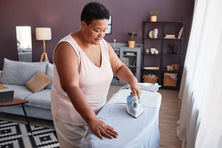 High angle view of mature black woman ironing clothes at home while caring for householdの写真素材