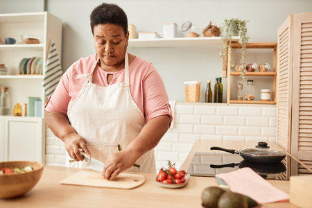 Warm toned waist up portrait of black senior woman cutting vegetables while making salad in cozy kitchen, copy spaceの写真素材