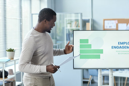 Side view portrait of black man giving presentation in office standing by digital screen and wearing...
