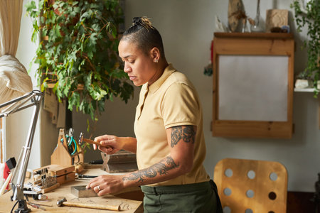Side view portrait of tattooed female artist creating handmade jewelry in cozy workshop lit by sunlightの写真素材