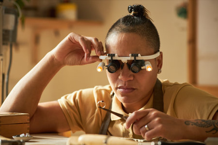 Female jeweler holding handmade ring and wearing magnifying goggles in cozy studioの写真素材
