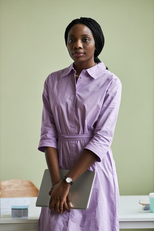 Vertical portrait of young black woman wearing purple outfit looking at camera against pastel green wall in officeの写真素材