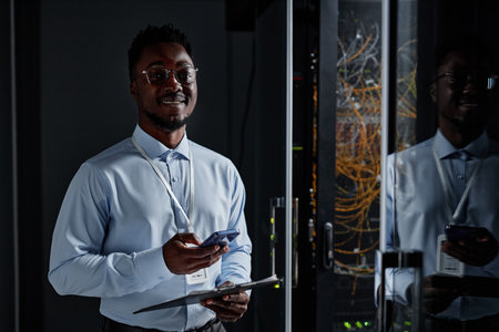 Waist up portrait of smiling black man as network engineer standing by server cabinet in data center...