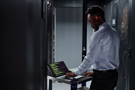 Side view portrait of black man as network engineer using laptop while setting up servers in data...