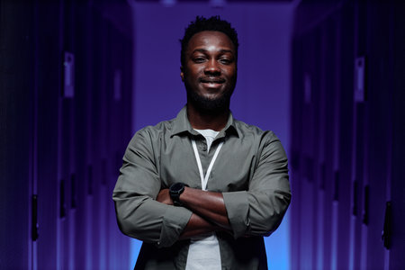 Waist up portrait of black young man standing in server room with arms crossed and looking at camera lit by purple neon lightの写真素材