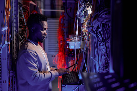 Side view portrait system administrator using computer in server room lit by neon light