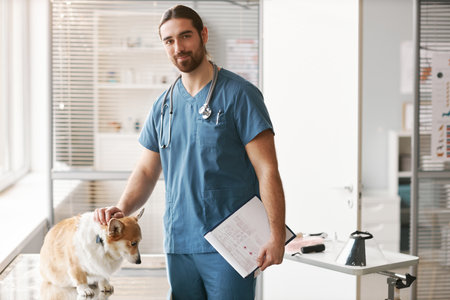 Young confident male veterinarian in blue uniform standing by workplace in vet clinics, cuddling...