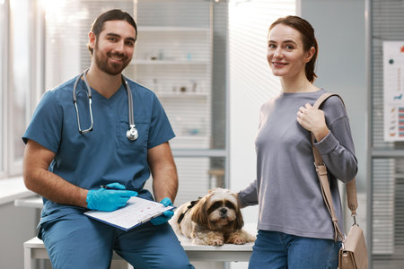 Young female owner of cute yorkshire terrier, her pet and veterinarinary clinician in blue uniform looking at camera in medical officeの写真素材