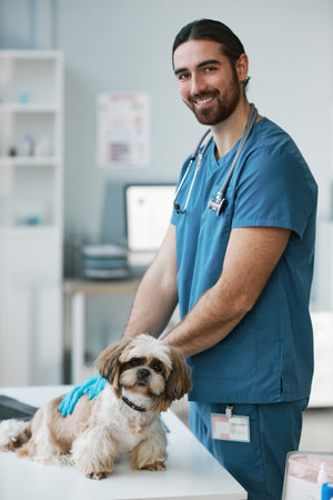 Young male veterinary doctor in uniform and gloves standing by medical table with yorkshire terrier and looking at camera during check upの写真素材