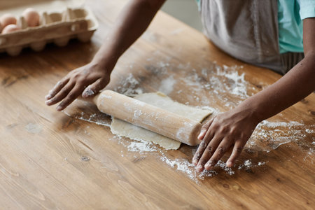 Close up of black little girl baking homemade pastry and rolling dough on dark wood kitchen counter,...