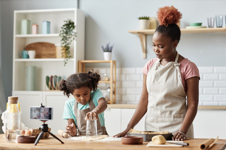 Waist up portrait of mother and daughter baking together in kitchen and recording video or...