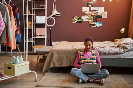Wide angle view of black teenage girl sitting on floor in pink room interior and using laptop, copy...