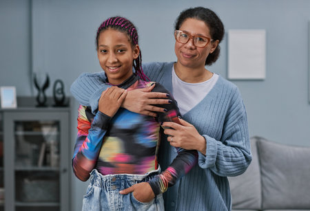 Waist up portrait of black mature woman embracing teenage daughter in minimal home interior, both...