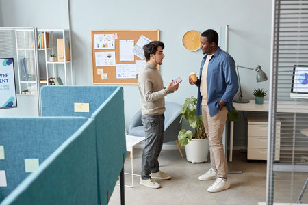 Full length side view portrait of two men wearing casual clothes chatting in office setting
