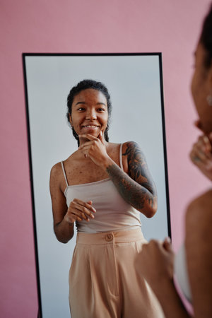 Vertical portrait of young black woman with tattoos and acne scars looking in mirror smiling confidentlyの写真素材