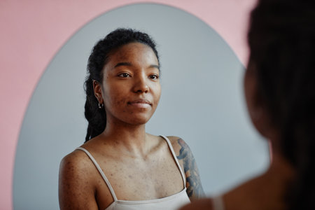 Young black woman with tattoos and acne scars looking in mirror smilingの写真素材