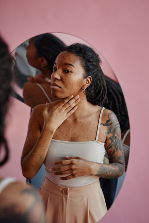 Vertical portrait of black young woman with acne scars looking in mirror insecure in appearanceの写真素材