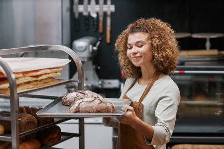 Positive small bakery owner with curly hair looking at tray with bread she made for sellingの写真素材