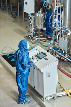Vertical portrait of worker in protective gear operating equipment at factory standing by control...