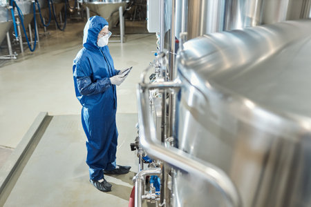 Side view at female worker wearing protective suit standing by steel tanks at industrial chemical...