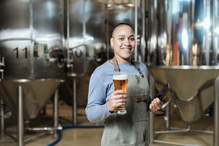 Waist up portrait of smiling female brewmaster holding beer glass and looking at camera while working at brewing factoryの写真素材