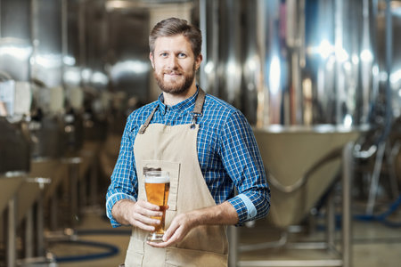 Waist up portrait of smiling bearded man holding beer glass and looking at camera while working at brewing factoryの写真素材