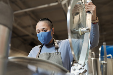 Low angle portrait of female brewmaster wearing mask opening tank at beer making factoryの写真素材