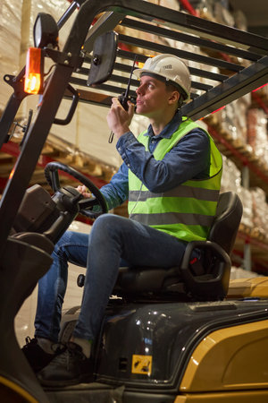 Vertical portrait of young male worker driving forklift truck in warehouse and speaking to portable radioの写真素材