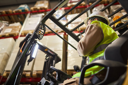 Low angle view of female worker wearing hardhat while operating forklift truck in warehouse and talking to portable radioの写真素材
