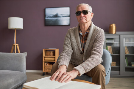 Blind senior man in eyeglasses reading a book in Braille at table in the living room