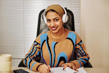 Front view portrait of young Middle Eastern woman wearing headphones in office and smiling at cameraの写真素材