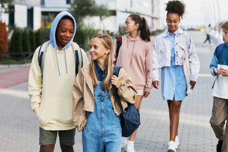 Cute intercultural school learners with backpacks chatting on their way home after classes while...