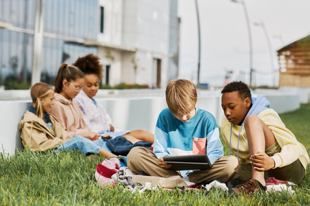 Two intercultural schoolboys looking at tablet screen while sitting on green lawn in park against...