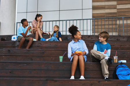 Two happy intercultural schoolmates sitting on wooden staircase at lunch break, having chat and eating sandwiches against group of kidsの写真素材