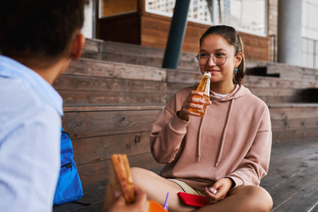 Happy schoolgirl in hoodie and eyeglasses drinking soda from bottle and looking at her friend during...