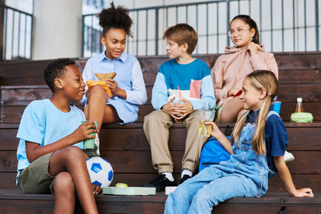 Youthful intercultural school friends with soda looking at one another while sitting on wooden staircase and chatting against other kidsの写真素材