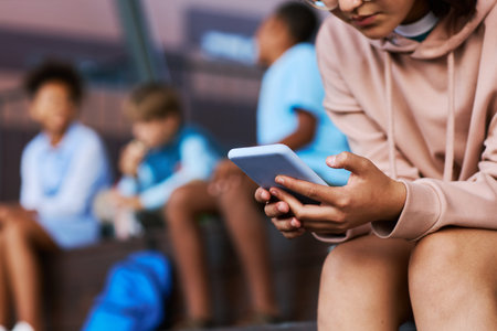 Close-up of secondary school learner with mobile phone watching online video while sitting against group of intercultural friendsの写真素材