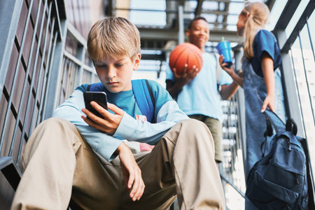 Adolescent schoolboy with backpack scrolling in smartphone while sitting on playground facilities...