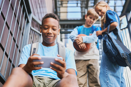 Happy African American schoolboy looking at screen of smartphone while watching online video against...