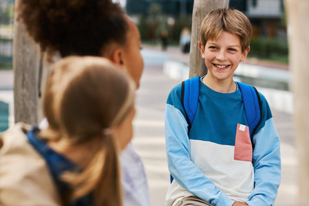 Cute laughing schoolboy in blue pullover chatting with two intercultural classmates after lessons...