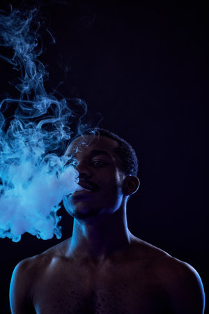 Young man standing in front of camera in darkness and releasing vapor out of his mouth during...