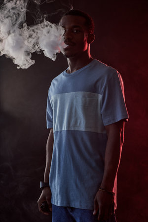 Young African American man in t-shirt and jeans holding electronic cigarette while standing in front...