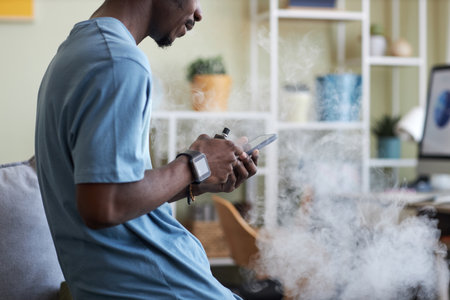 Side view of young black man in t-shirt standing in front of camera in living room and texting in smartphone while smoking e-cigaretteの写真素材
