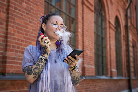Young woman with tattoos on arms and hands smoking electronic cigarette and scrolling in mobile...