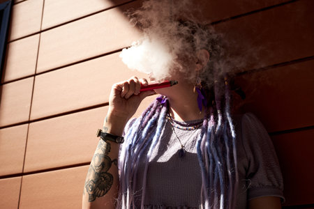 Hidden face of young woman with dreadlocks holding e-cigarette while standing against wall and blowing cloud of smoke out of mouthの写真素材