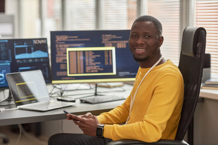 Portrait of smiling black man looking at camera while sitting at workplace in software development...