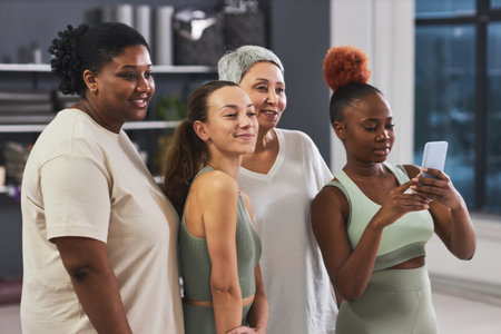 Group of multiethnic women posing on camera of smartphone together standing in gymの写真素材