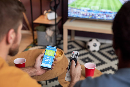 Close up of two men watching football match on TV and holding smartphone with online sports betsの写真素材